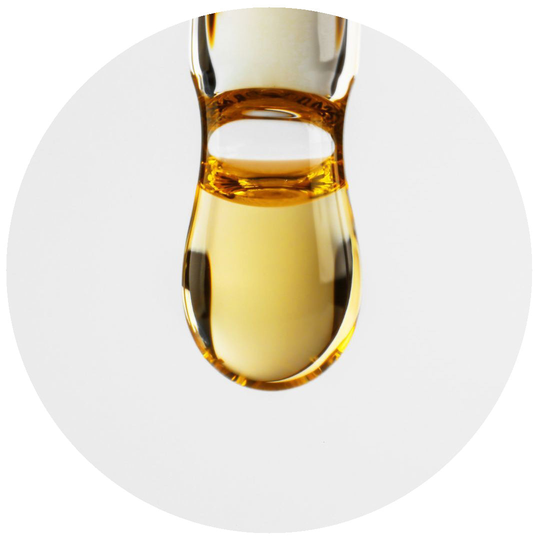 A close-up of a golden-yellow liquid drop suspended from the tip of a glass dropper against a plain white background.