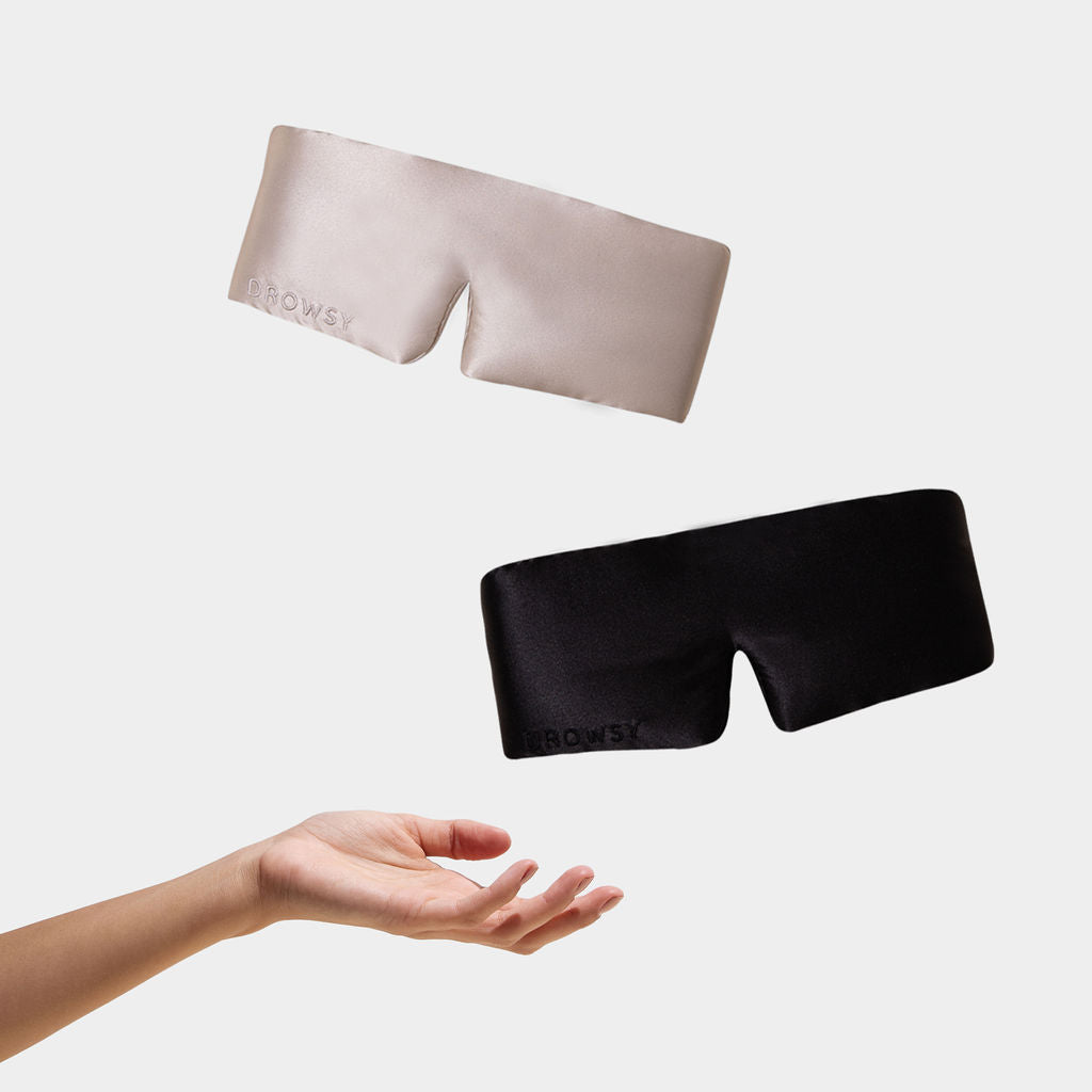 Two luxury silk eye masks from Drowsy’s Lovers Pack in beige and black float above an outstretched hand, each embossed with DROWSY—perfect for beauty sleep against a clean white background.