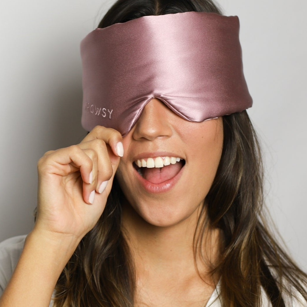 A smiling woman with long brown hair wears a shiny mauve sleep mask, lifting it slightly with her right hand, revealing one eye. She appears cheerful against a plain light background.