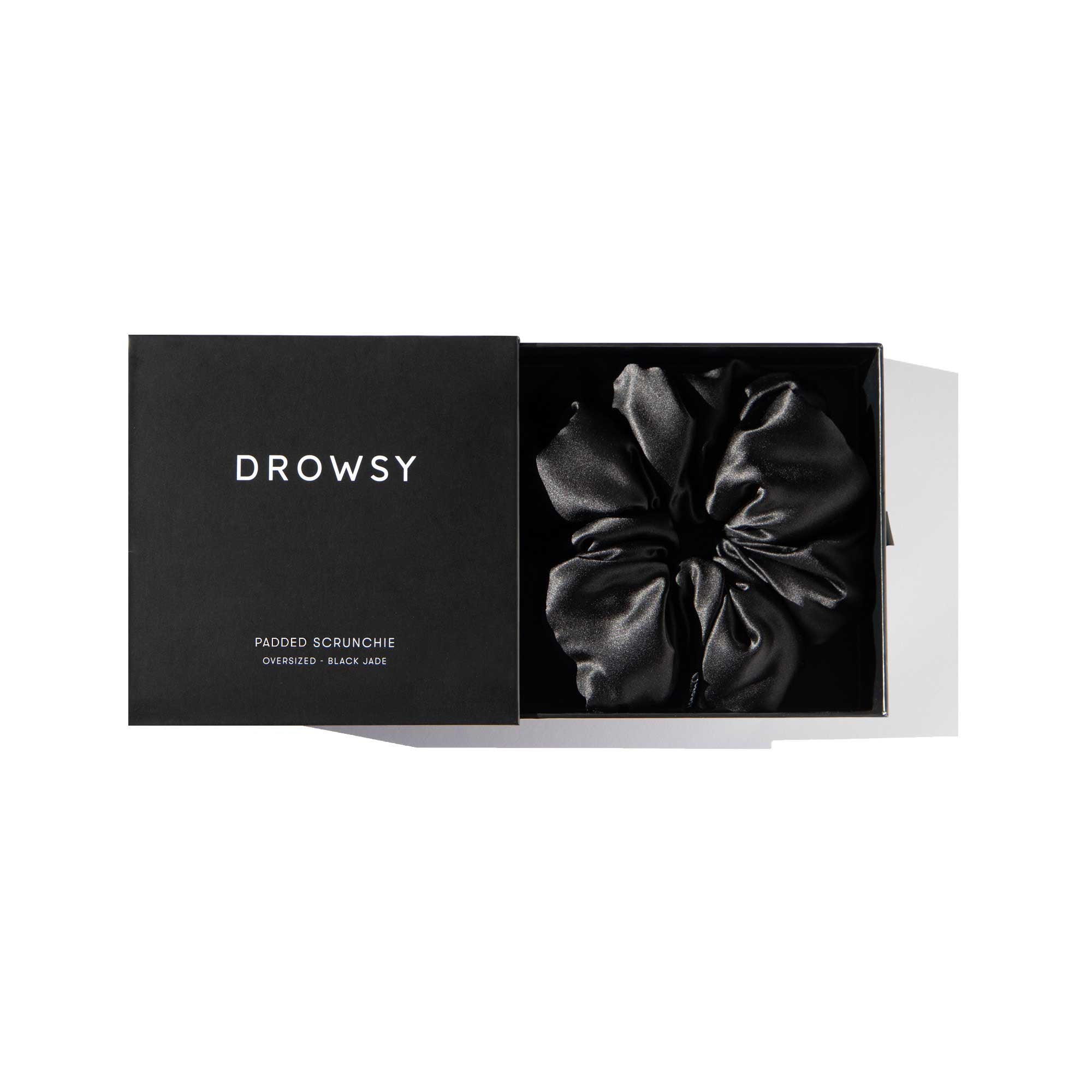 A black gift box labeled us.drowsysleepco with the lid partially open reveals the Black Jade - Padded Scrunchie inside, crafted from padded silk. Text on the box reads: Padded Scrunchie Oversized Black 100%. Perfect for hair health.