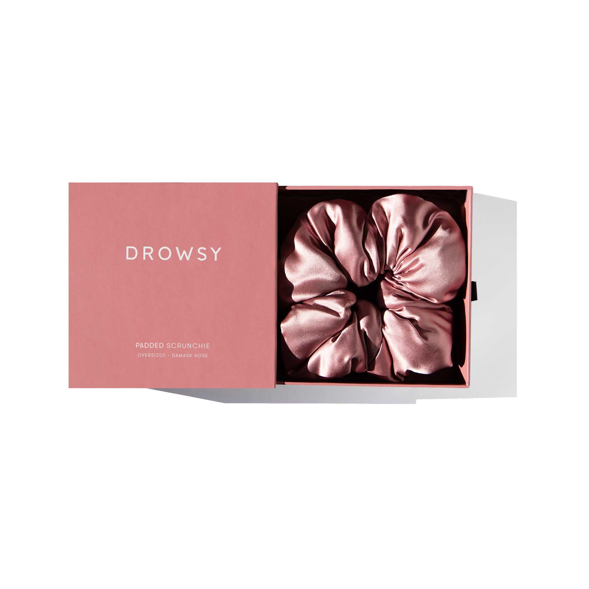 A pink box labeled us.drowsysleepco holds the Damask Rose - Padded Scrunchie, a shiny, oversized silk hair accessory neatly coiled inside. The partially open lid reveals it against a white background, ideal for elevating your hair health.