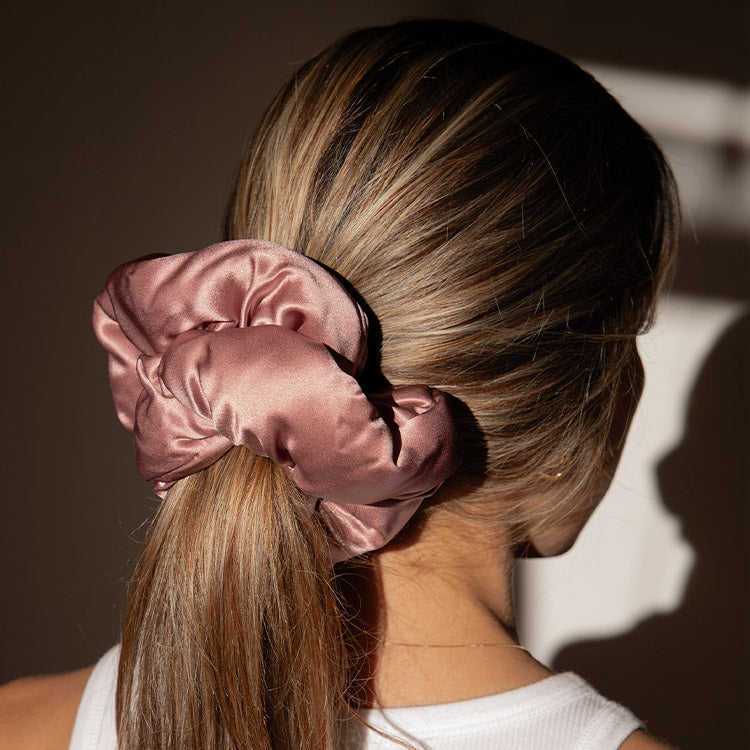 A woman with light brown hair in a low ponytail wears a shiny mauve scrunchie, dressed in white. Soft shadows evoke damask rose, reflecting the gentle elegance of the drowsysleepco Sensual Sleep Bundle - Damask Rose.