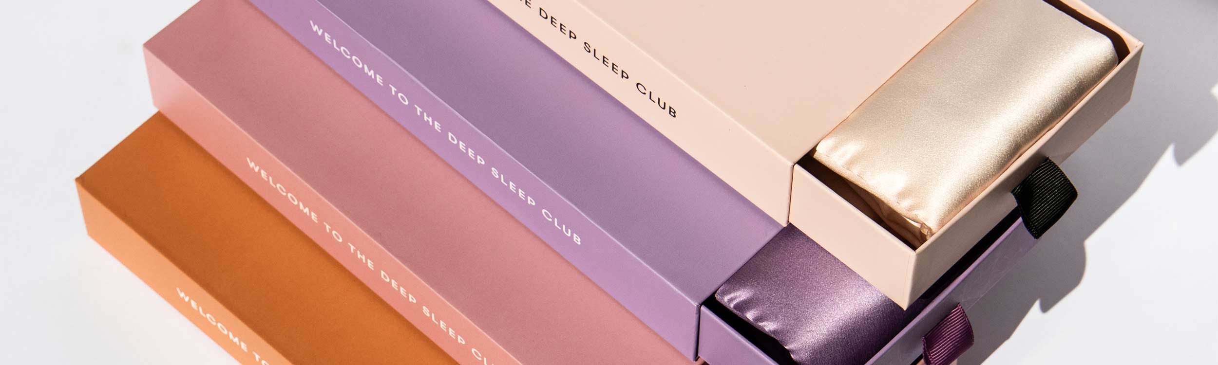 Four rectangular boxes in shades of peach, lavender, purple, and tan, each labeled WELCOME TO THE DEEP SLEEP CLUB, with satin eye masks in matching colors partially visible inside the open boxes.