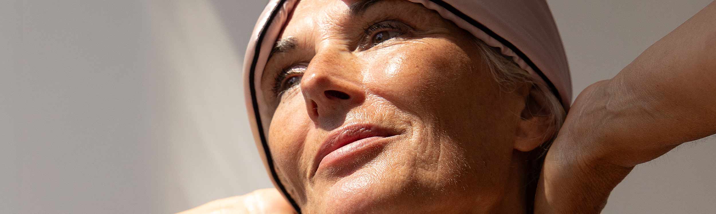 A close-up of an older woman wearing a light pink headscarf, looking upward with a serene expression, sunlight highlighting her face and smooth skin.