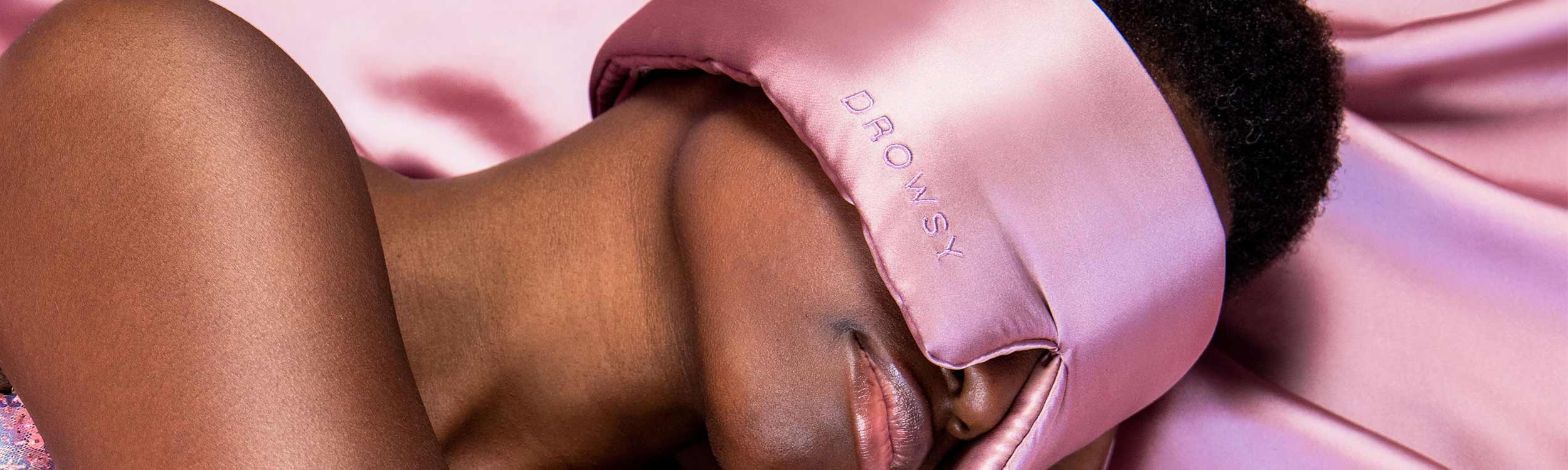 A person with short hair lies on pink satin fabric, wearing a large silky pink sleep mask embroidered with the word DROWSY.