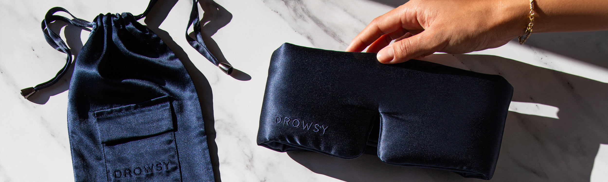 A hand holds a dark blue satin sleep mask next to its matching drawstring pouch on a white marble surface. The brand name DROWSY is visible on both the mask and pouch.