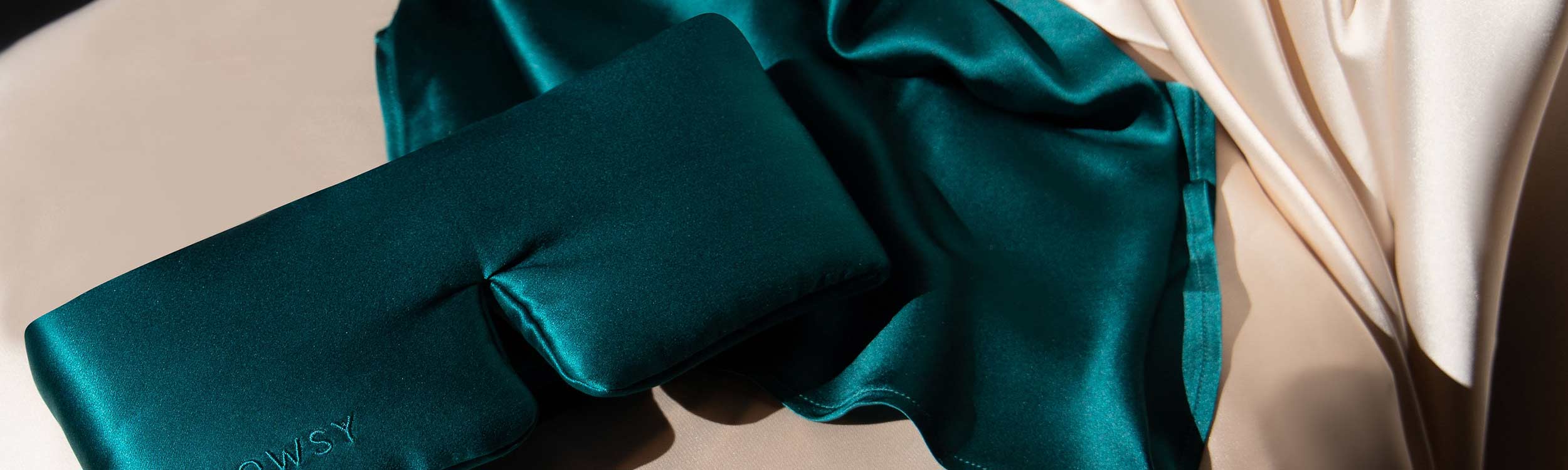 A teal silk eye mask and matching silk pillowcase rest on a beige surface, illuminated by sunlight, with a cream-colored silk fabric draped in the background.
