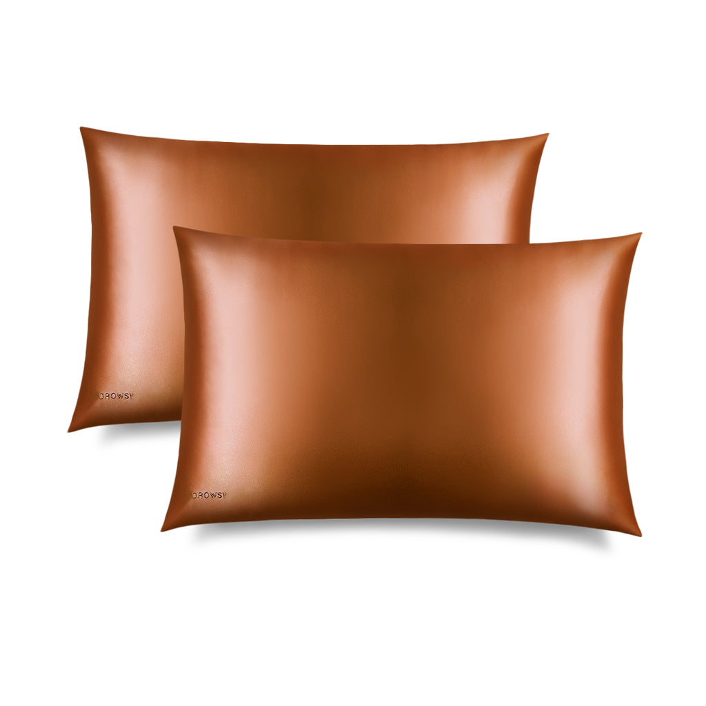 Two shiny brown us.drowsysleepco Pumpkin Spice Silk Pillowcases are shown slightly overlapping on a white background. A skincare and beauty essential, they protect hair and feature a small logo in the bottom corner.