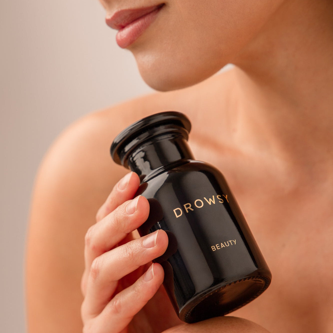 A person with bare shoulders holds a glossy black Apothecary Glass Storage Jar from drowsysleepco near their chin, with the image focusing on their lower face, hand, and the sleek branded jar.