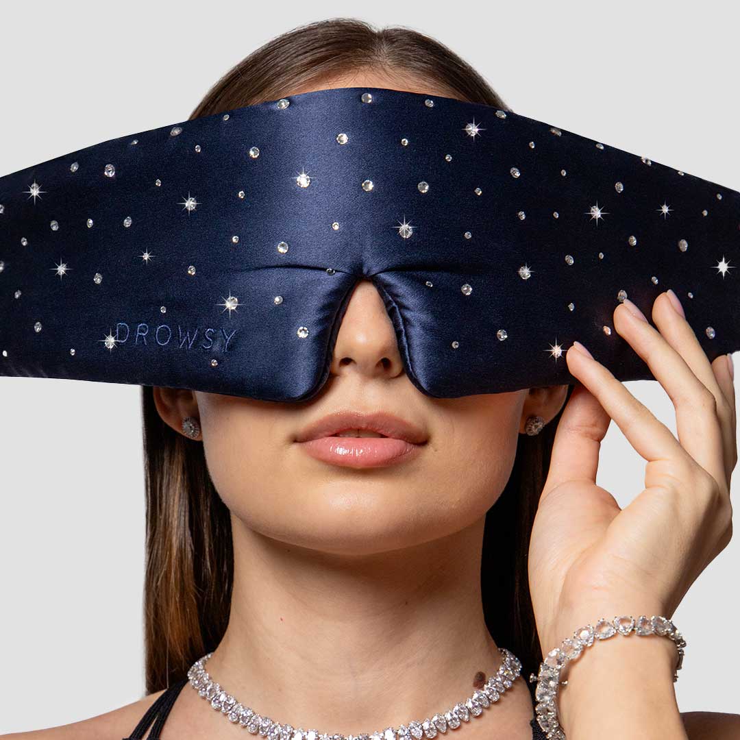 A woman wearing a wide, dark blue, star-studded sleep mask labeled “DROWSY,” holding it with one hand. She is accessorized with sparkly jewelry and pictured against a plain light background.
