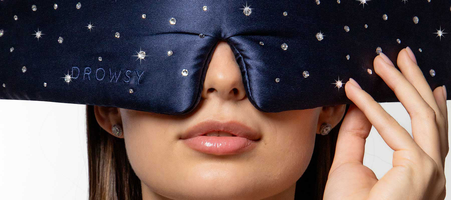 A woman holds a dark blue, gem-studded sleep mask embroidered with DROWSY over her eyes. Her lips and lower face are visible, and the background is white.