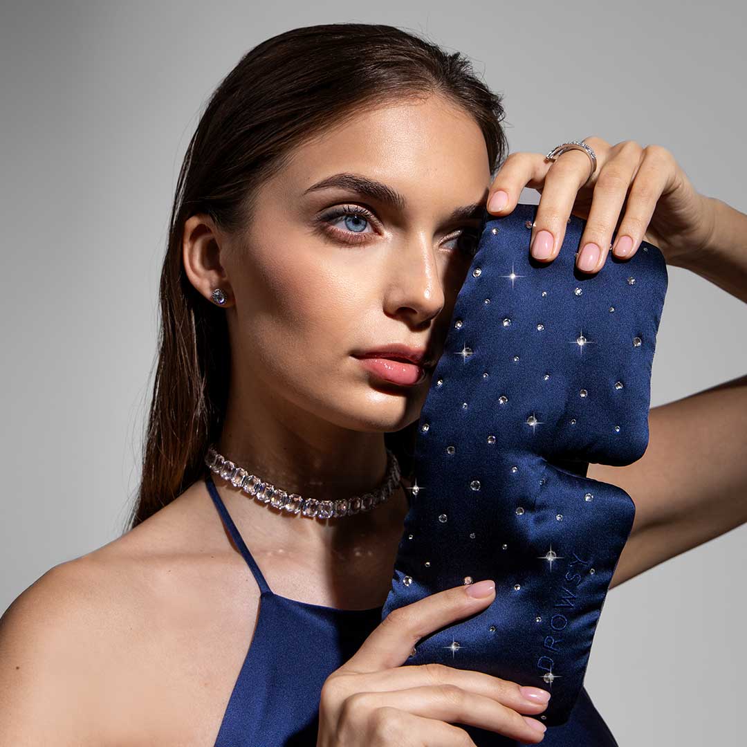 A woman with long brown hair holds a dark blue, jeweled sleep mask beside her face. She wears a sparkly choker necklace, earrings, a ring, and a navy satin outfit, posing against a neutral background.