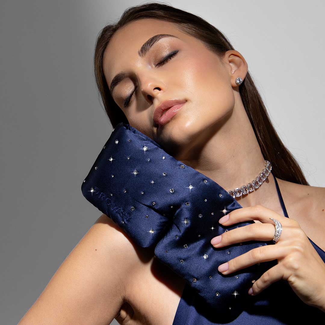 A woman with long brown hair wears a navy blue dress and jewelry, and gently rests a navy blue, embellished pillow against her face with her eyes closed, looking serene and relaxed.