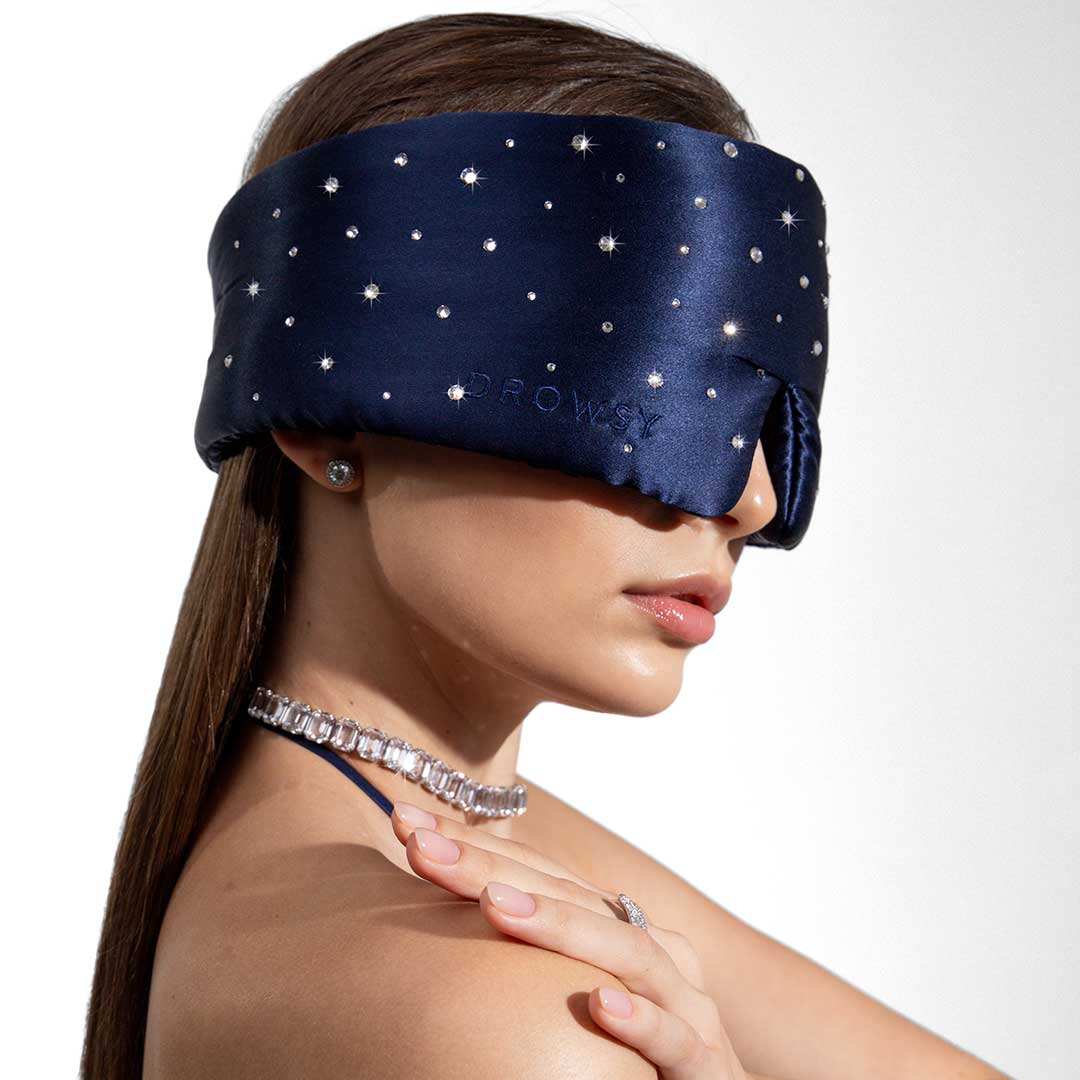 A woman with straight brown hair wears a dark blue, star-patterned silk sleep mask. She has stud earrings and a sparkling necklace, and she gently touches her shoulder with one hand. The background is white.