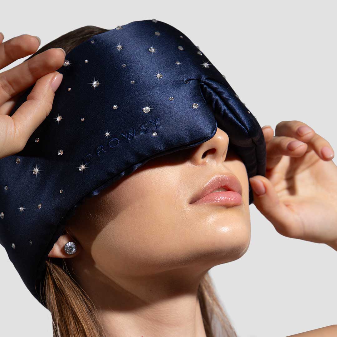 A woman wears a dark blue, star-embellished sleep mask, holding it in place with both hands. Her face is partially visible with glossy lips, and she has stud earrings. The background is plain and light-colored.