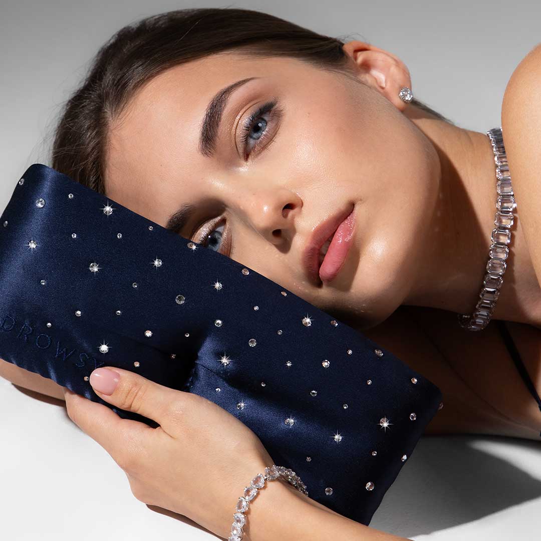 A woman with blue eyes and light makeup rests her head on a navy blue, rhinestone-studded pillow. She wears a sparkly necklace, bracelet, and earrings, with a soft and serene expression on her face.