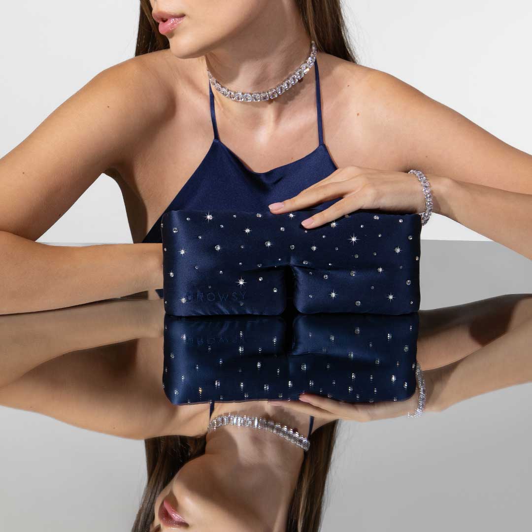 A woman in a navy blue halter dress adorned with a rhinestone choker and bracelet sits at a reflective table, holding a navy clutch decorated with small sparkling gems and stars. Her face is partially out of the frame.