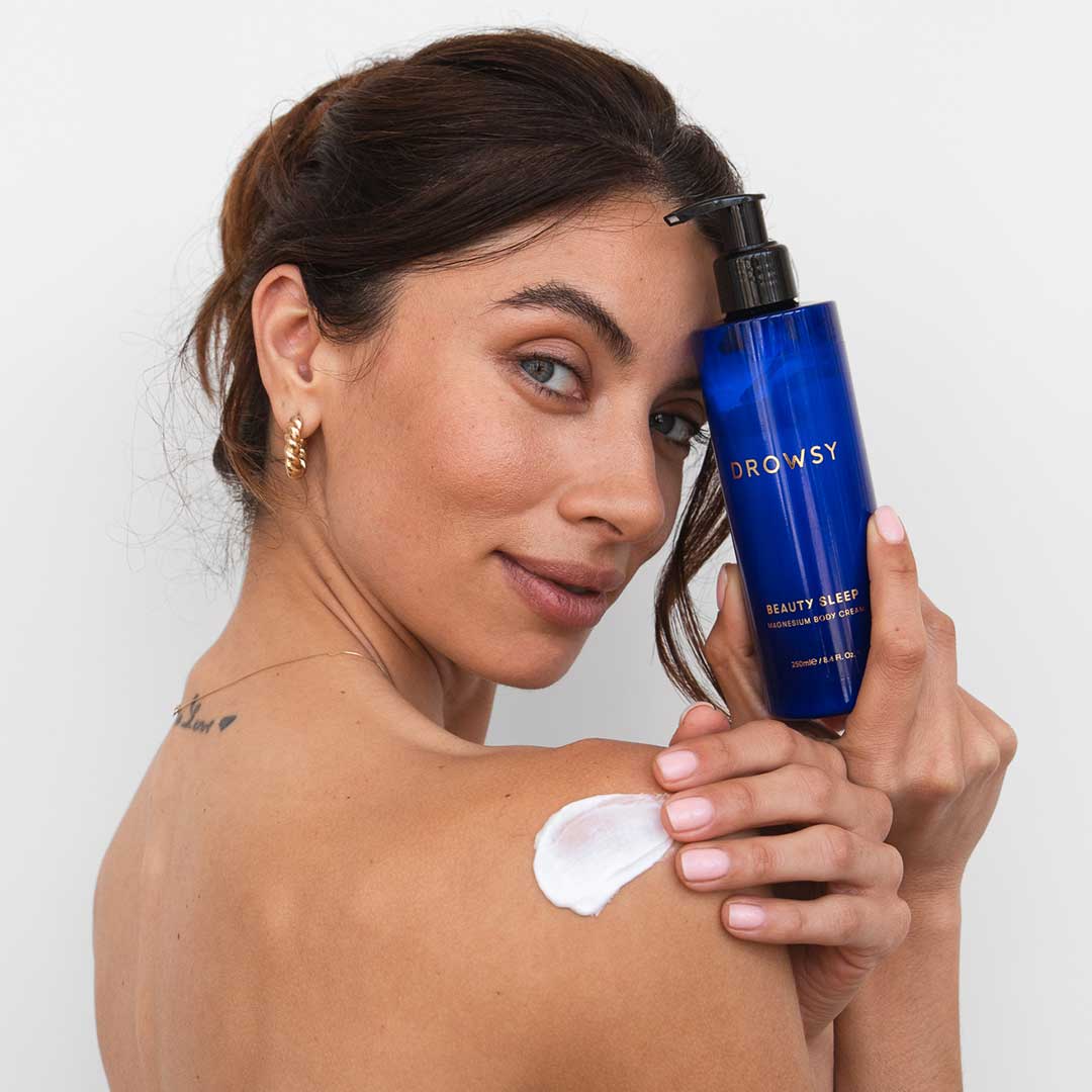 A woman with brown hair and gold hoop earrings applies white lotion to her shoulder from a blue bottle of us.drowsysleepco’s Beauty Sleep Body Cream. She smiles slightly at the camera.