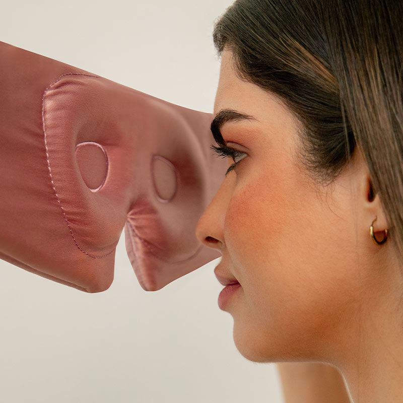 A woman faces a strange padded mask-like object with two eye holes, positioned closely in front of her eyes. She has straight brown hair, light makeup, and wears a small gold hoop earring. The background is plain and neutral.