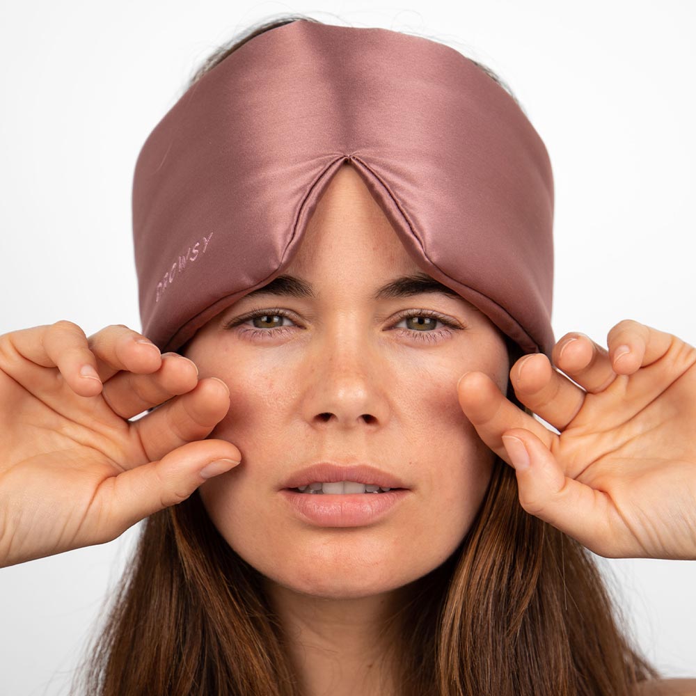 A woman gently touches the Damask Rose silky mauve blackout sleep mask by us.drowsysleepco on her forehead while looking at the camera.