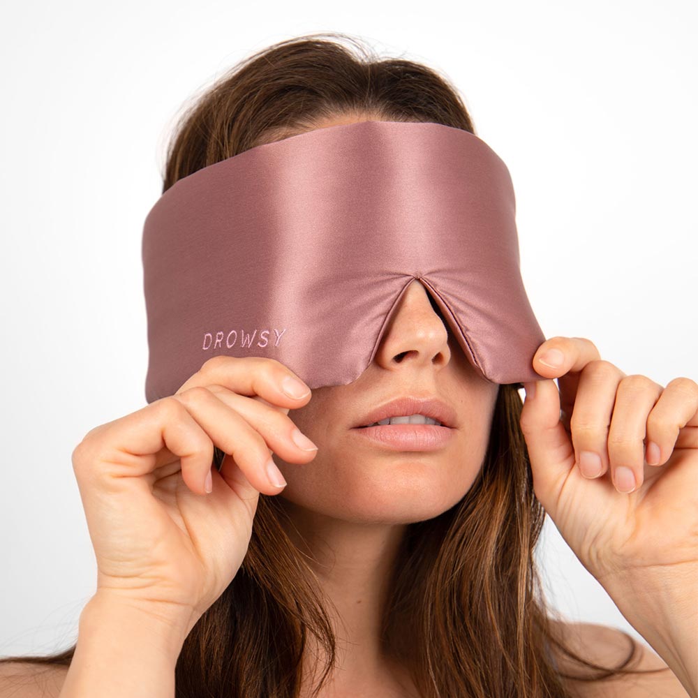 A woman adjusts a mauve silk sleep mask labeled Drowsy, covering her eyes, against a plain white background.