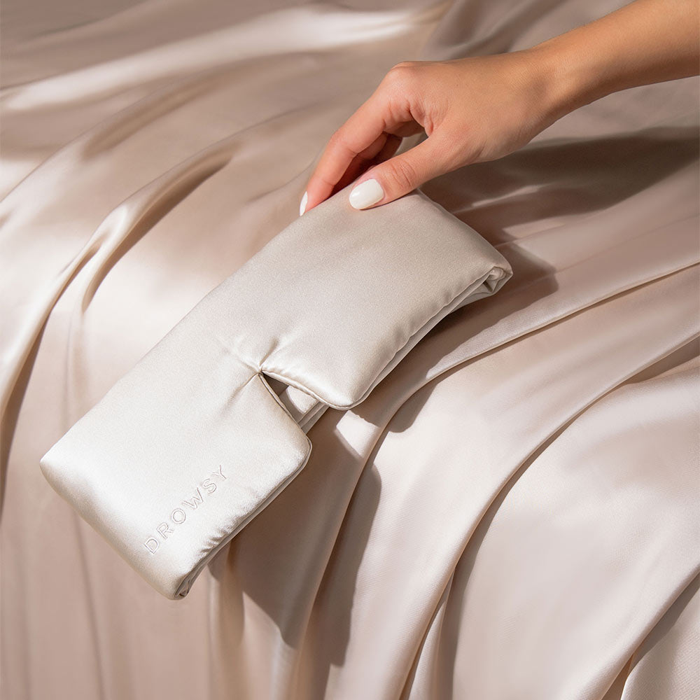 A hand holds a Dusty Gold silk eye mask by us.drowsysleepco with a blackout design over matching silk sheets. DROWSY is embroidered on the mask, evoking luxury and relaxation.