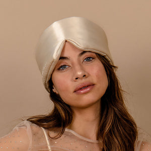 A woman with long brown hair wears the Dusty Gold silk turban by us.drowsysleepco and a sheer, embellished beige top, posing against a plain beige background—evoking the luxurious vibe of a signature Drowsy silk eye mask.