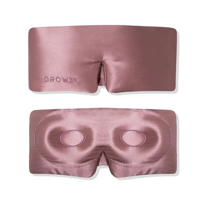 A pink silk sleep mask shown from the front and back; the front is smooth with DROWSY embroidered, while the back has eye contours and an opening for the nose.