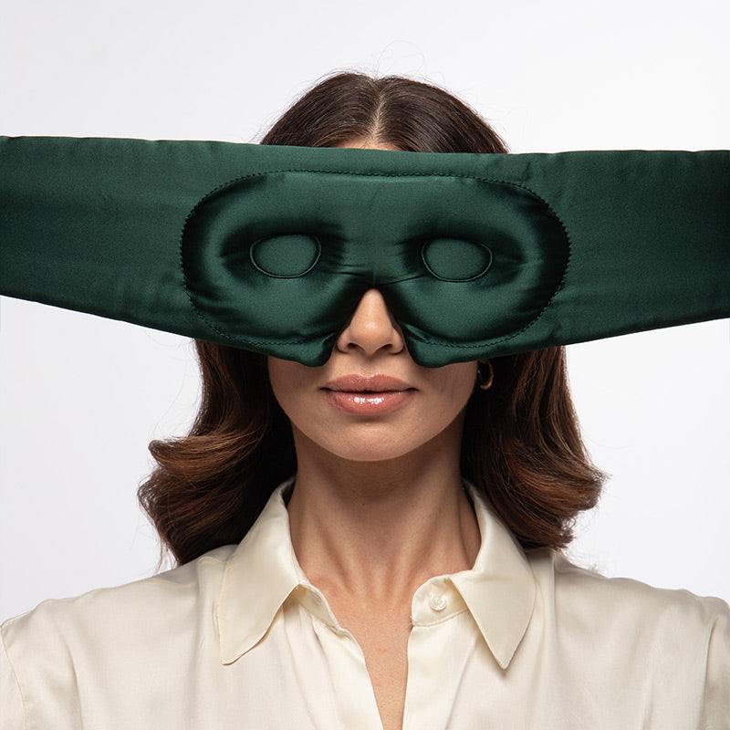 A woman faces forward, wearing the us.drowsysleepco Ivy Green - Eyelash Protecting Mask over her eyes and head. The dark green satin silk mask features special openings to protect her eyelashes while maintaining an elegant look. She is dressed in a white blouse against a plain white background.