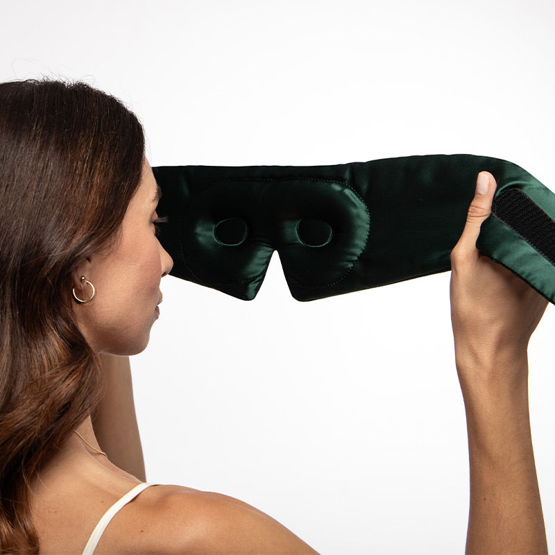 A woman with brown hair holds the us.drowsysleepco Ivy Green - Eyelash Protecting Mask, featuring a dark green contoured design with eye cutouts, as she prepares to put it on against a plain white background.