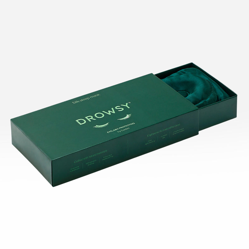 An Ivy Green - Eyelash Protecting Mask by us.drowsysleepco, featuring contoured eyecups and crafted from silk, is partially visible inside an open dark green box labeled “Drowsy” with product information displayed on the lid.