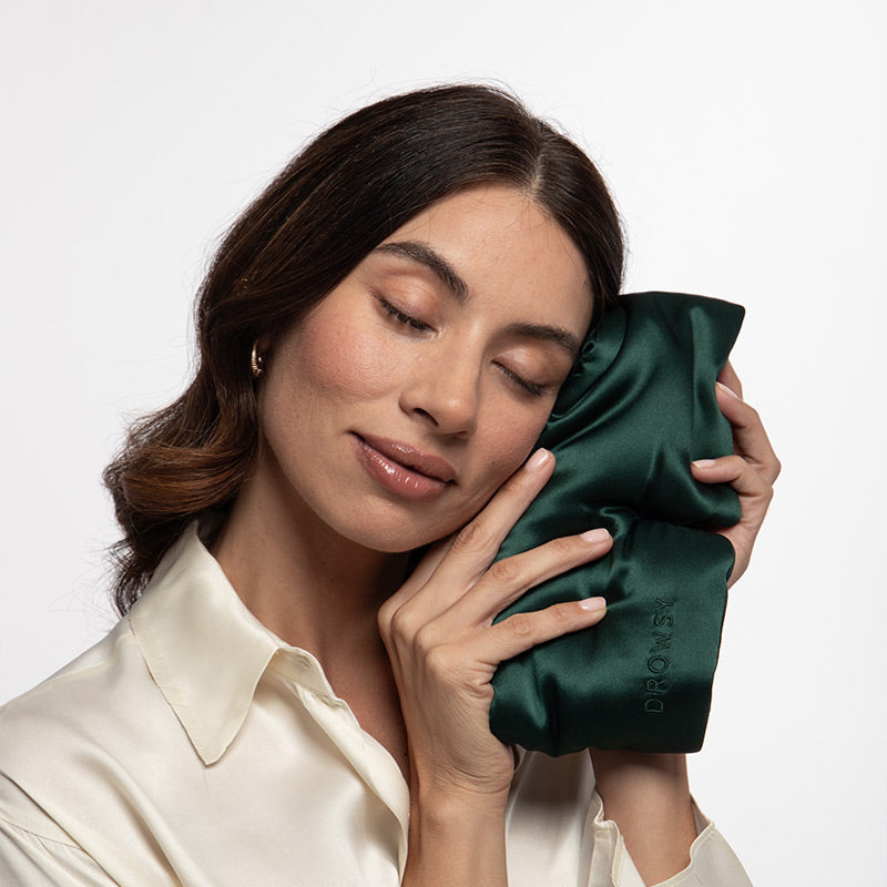 A woman with long brown hair in a cream blouse gently holds and rests her face on a dark green, silky pillow, exuding calm and contentment against a white background—her tranquility elevated by the us.drowsysleepco Ivy Green - Eyelash Protecting Mask, designed to promise restful nights.