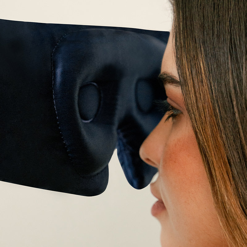 A close-up side view of a woman wearing a dark blue sleep mask, with her eye and part of her face visible, highlighting the masks cushioned eye area.