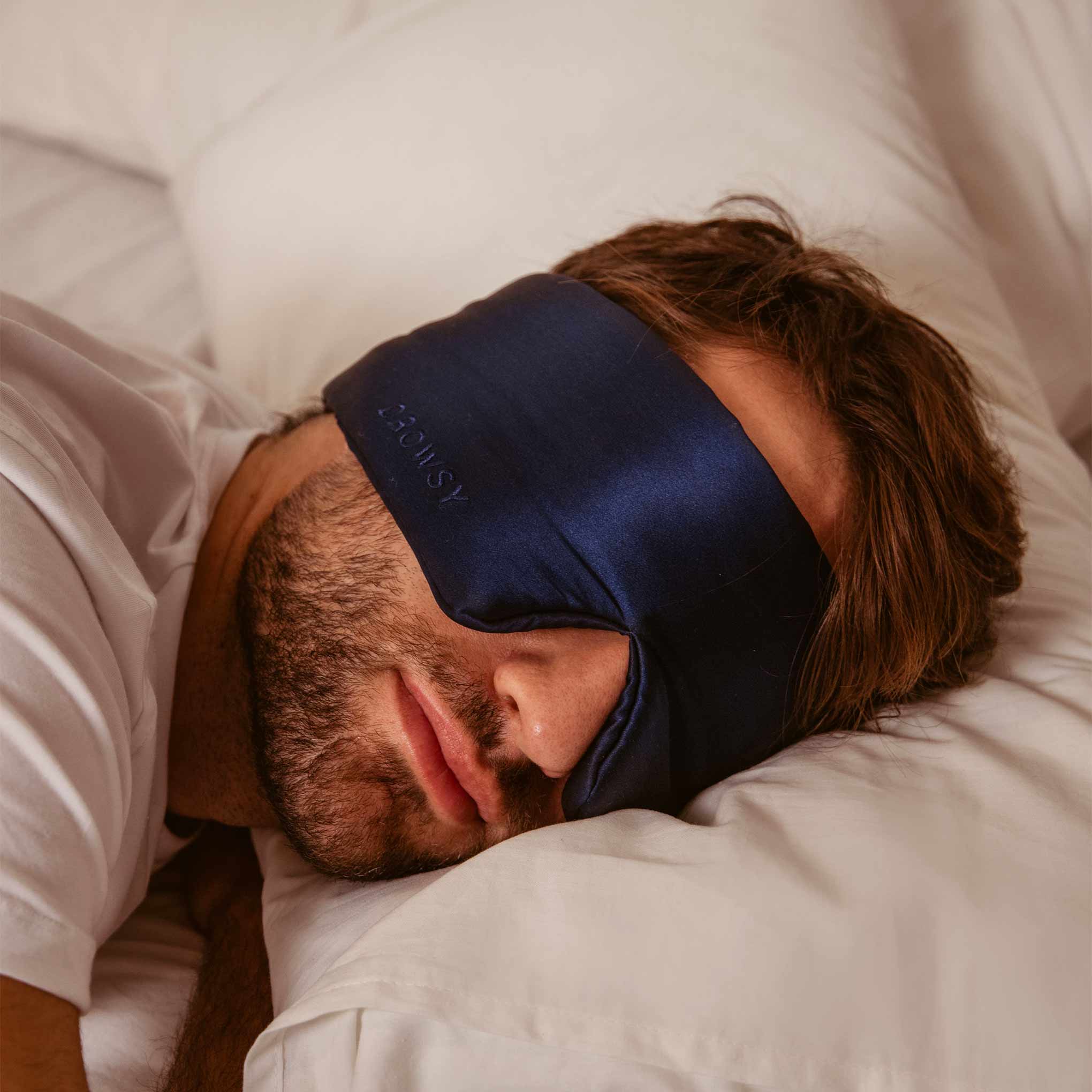 A person with short brown hair and facial hair, wearing a drowsysleepco Midnight Blue Sleep Mask from the 2-Pack Sleep Masks (Paris Love Story + Midnight Blue), rests on their side in bed with white pillows and a white shirt.
