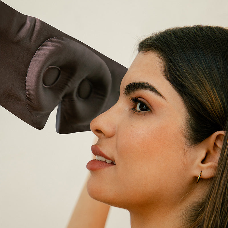A woman with long dark hair and hoop earrings is about to put on a dark satin sleep mask, holding it near her forehead while looking slightly upward and smiling softly.