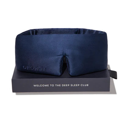 A dark blue silk sleep mask with “DROWSY” embroidered on the side, displayed on top of a gray box labeled “WELCOME TO THE DEEP SLEEP CLUB.”.