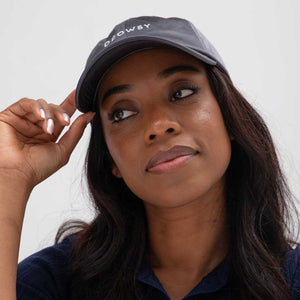 A woman with long dark hair wears the Drowsy Silk Cap - Moonlight Shadow by Drowsy, combining style and hair protection. She touches the brim thoughtfully against a plain light background.