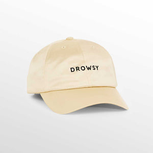 The Drowsy Silk Cap in Dusty Gold is a beige baseball cap with “DROWSY” in bold black letters on the front, shown against a light gray background.