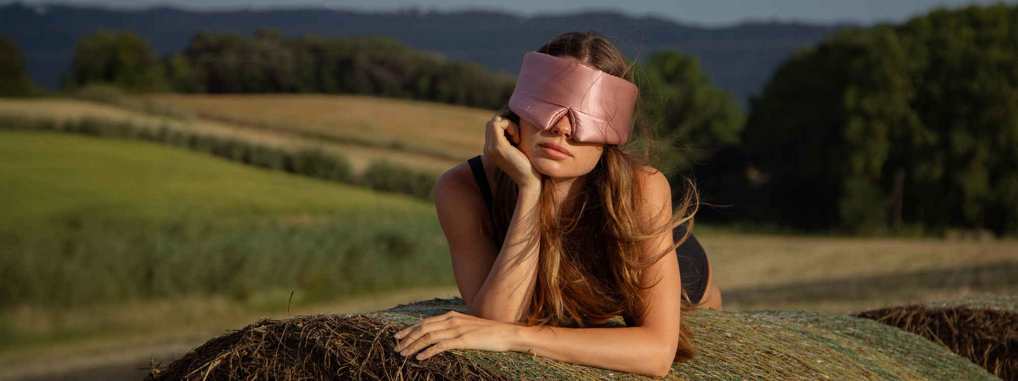 A woman wearing a pink sleep mask reclines on a hay bale in a rural field, surrounded by green grass and distant trees under a clear sky.