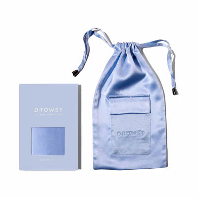 A pale blue Drowsy silk sleep mask pouch with a matching drawstring bag, both placed on a white background. The pouch has a clear window displaying the contents inside.