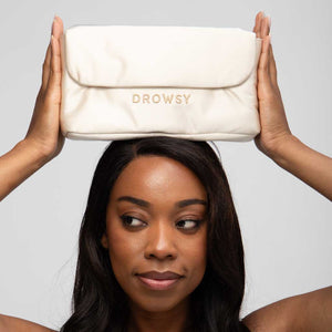 A woman with long dark hair holds the Drowsy Silk Necessaire in Dusty Gold above her head, gazing to the side. The simple, light background highlights this elegant Drowsy pouch as a chic addition to any beauty routine.