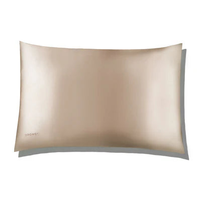 A rectangular champagne-colored silk pillowcase with a smooth, shiny texture and a small embroidered brand name in the lower left corner.