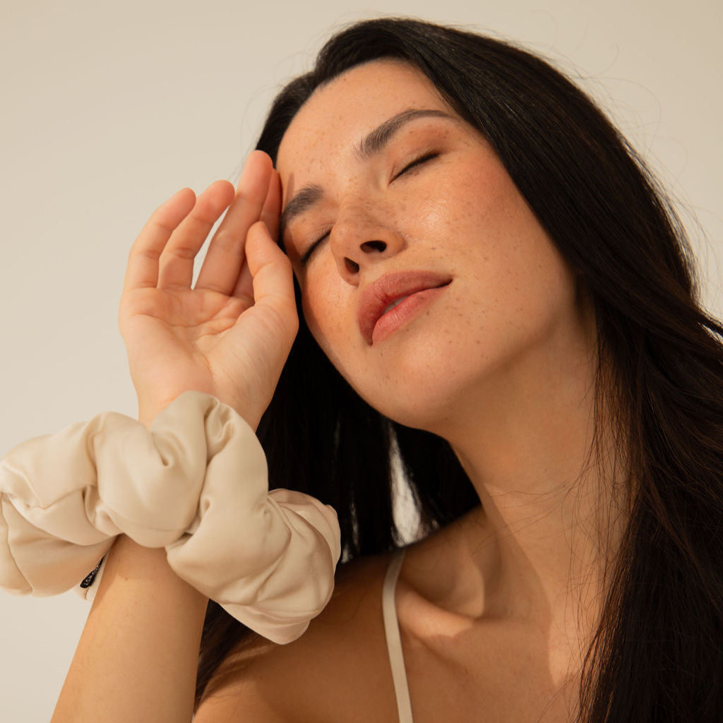 A woman with long dark hair closes her eyes and gently touches her forehead with her hand. She wears a cream-colored scrunchie on her wrist and a thin strap top, with soft lighting highlighting her serene expression.