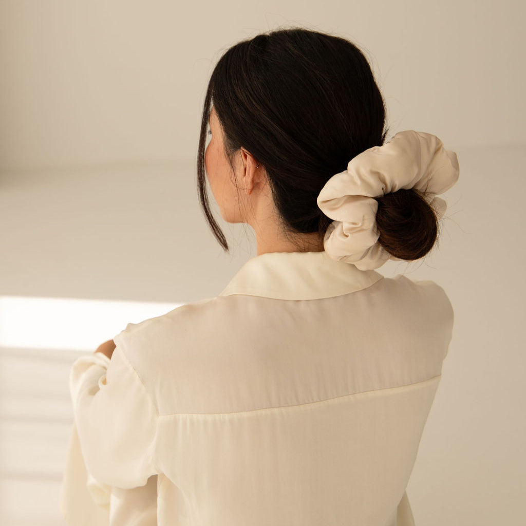 A woman with dark hair tied in a low bun with a large, beige scrunchie is seen from behind, wearing a light-colored blouse in a softly lit room.
