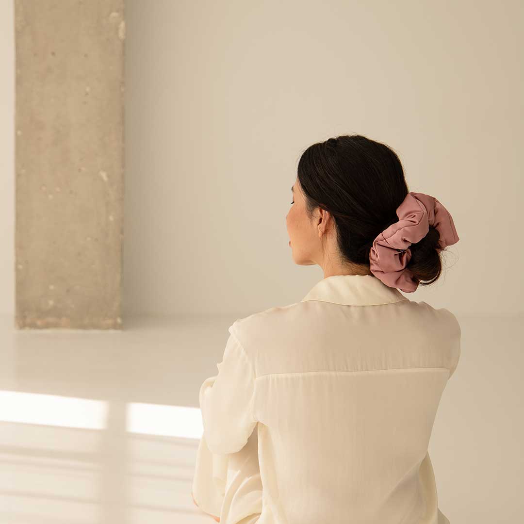 A woman with dark hair tied in a pink scrunchie sits on the floor facing away, wearing a cream-colored shirt in a minimal, sunlit room with light walls and a concrete pillar.