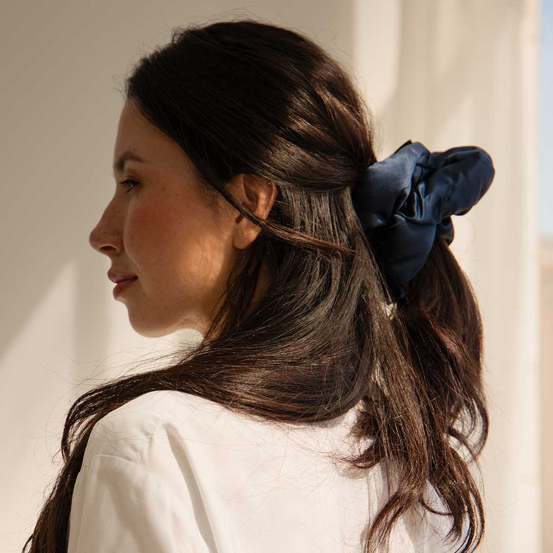 A woman with long brown hair, wearing a white top and the drowsysleepco Midnight Blue - Padded Scrunchie, gazes to the side in soft natural light.