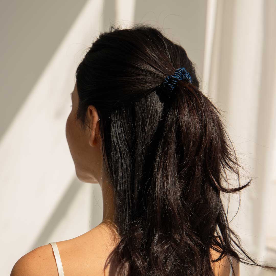 A woman with long, dark hair tied in a ponytail with a dark blue scrunchie stands facing away from the camera in bright natural light, wearing a white sleeveless top.