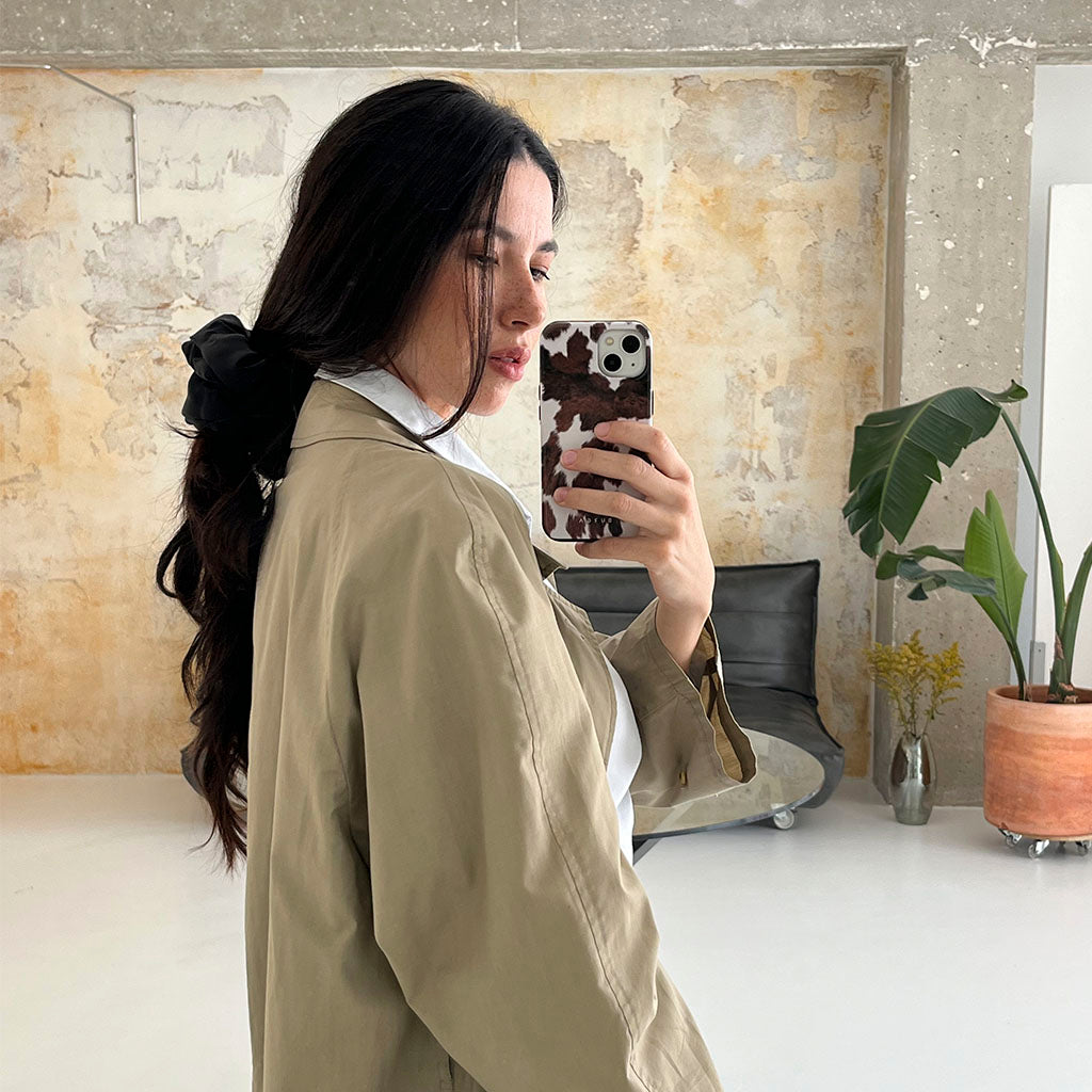 A woman with long dark hair tied with a black ribbon takes a mirror selfie. She wears a beige trench coat over a white shirt in a modern, minimal room with plants and a distressed wall.