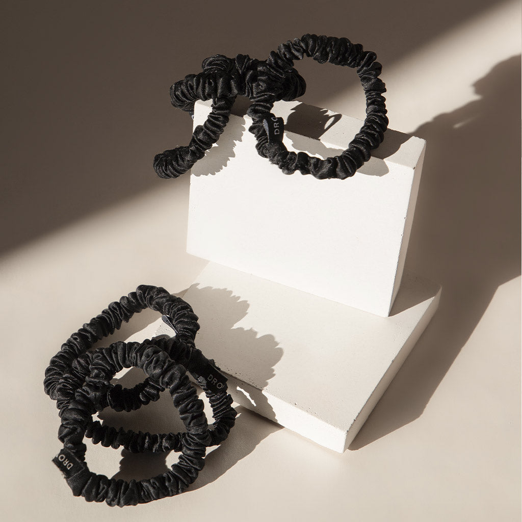 The us.drowsysleepco Skinny Scrunchie Set – Black Jade features three black silk scrunchies displayed on minimalist white blocks, casting soft shadows—a chic and healthy way to elevate your hair.