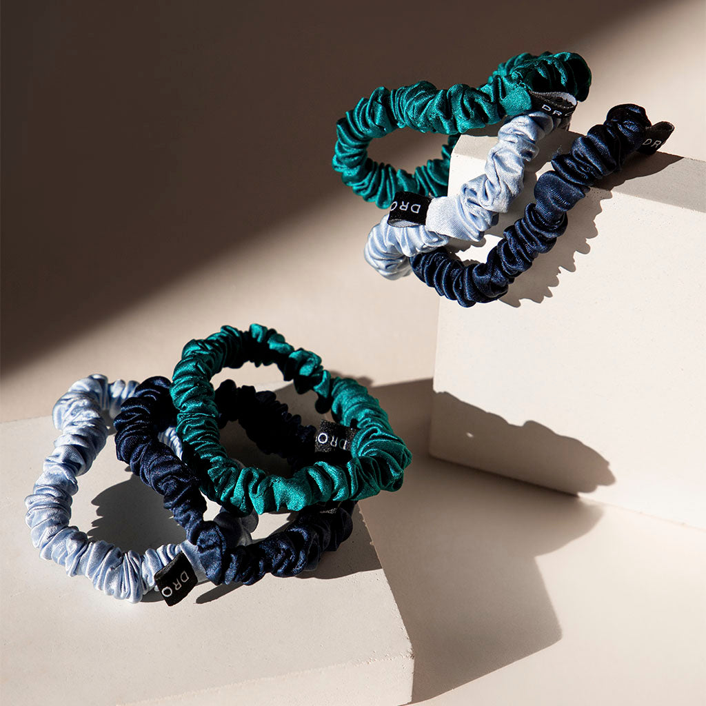 The us.drowsysleepco Skinny Scrunchie Set – Blue & Green Shades features three luxurious 22 momme silk scrunchies in green, navy, and light blue, elegantly displayed on beige geometric blocks with soft sunlight accents.